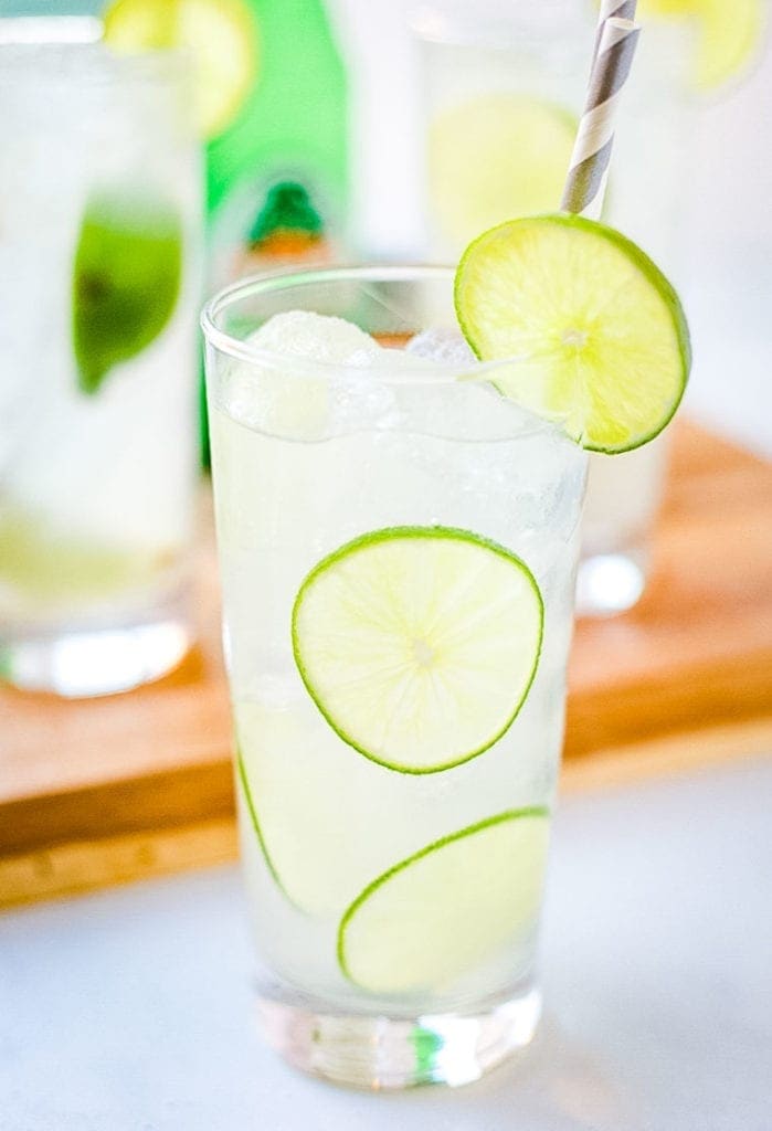 Close up of a Paloma Cocktail in high glass. Garnish with lime wedge on rim alone with slices in drink. Paper straw in drink that is gray and white striped.