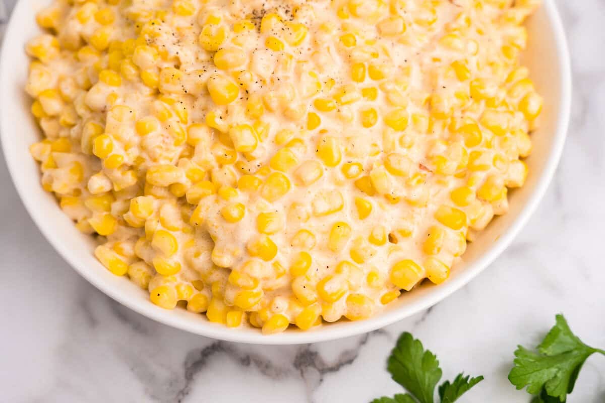 A white serving bowl filled with the finished creamed corn, garnished with cracked black pepper, ready to be served with fresh parsley nearby for garnish.