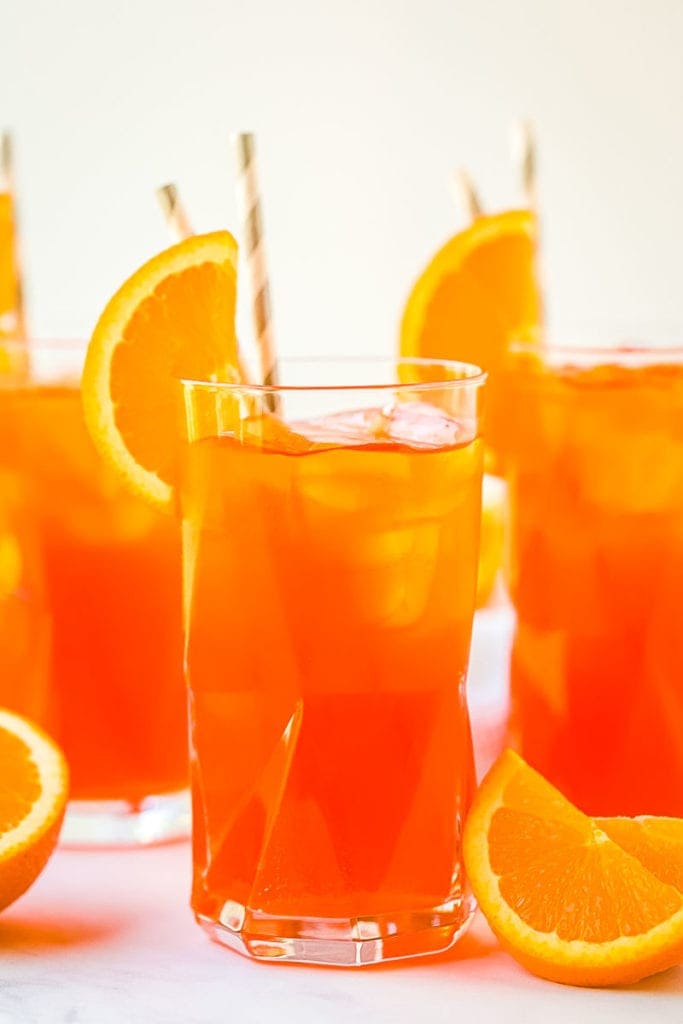 Close up image of an Aperol Spritz in tall glass with two in the background. Orange slices on marble surface next to glasses. 