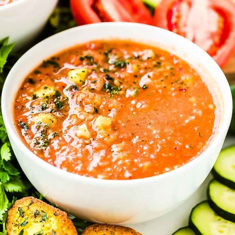 Gazpacho in white bowl with cucumber, tomato, and garlic toast around it