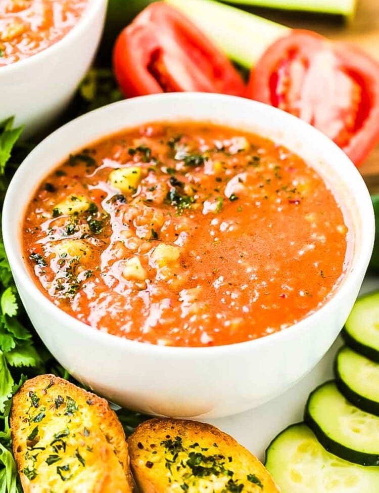 Gazpacho in white bowl with cucumber, tomato, and garlic toast around it