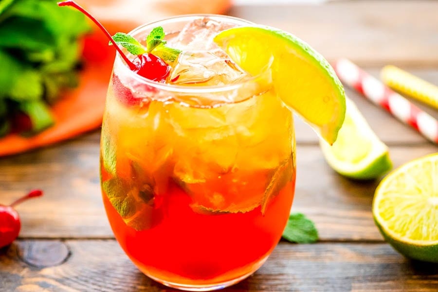 A glass filled with mai tai cocktail. Garnished with a lime wedge on rim, maraschino cherries and mint in the drink.