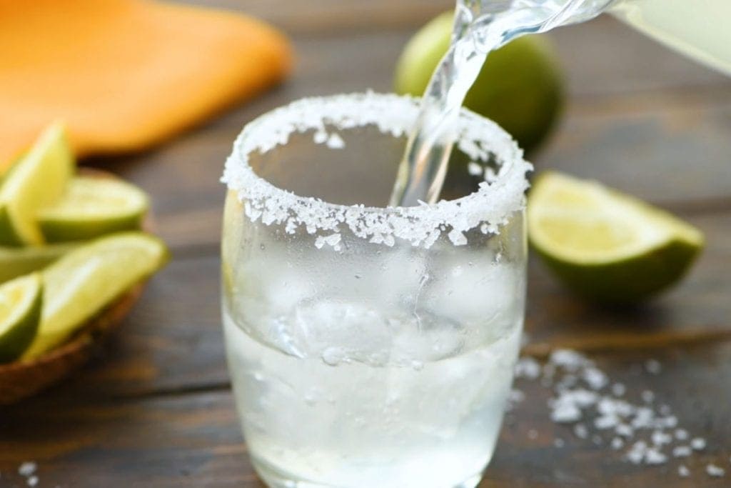 Pouring Margarita out of a glass pitcher and into a glass that's rimmed with salt.