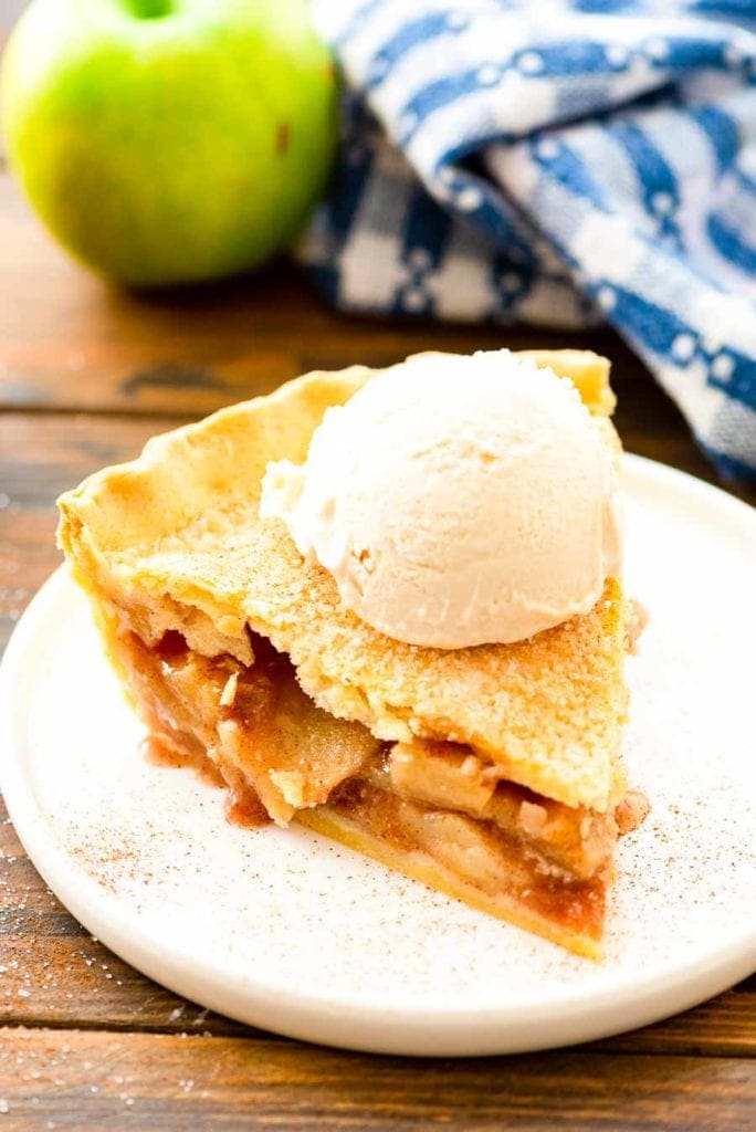 Slice of Apple Pie topped with ice cream on a white plate with blue and white napkin in background and a green apple.