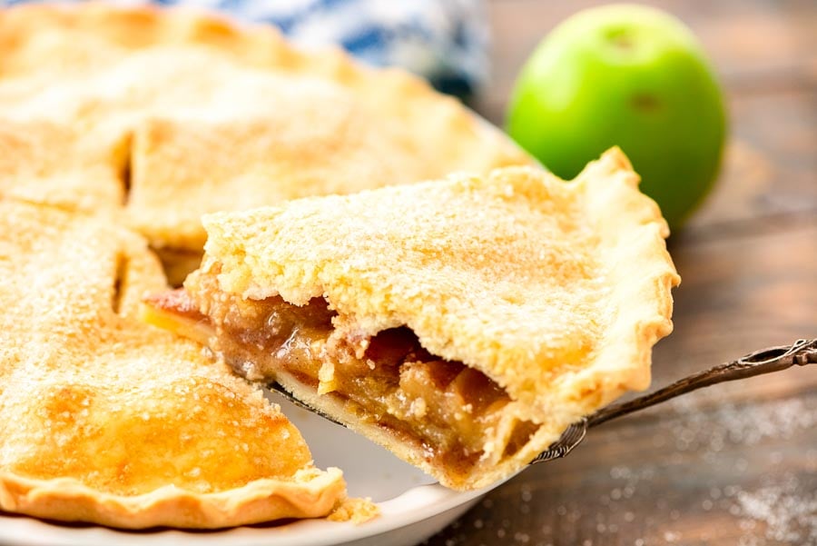 Apple Pie Slice on metal spatula being pulled out of a freshly baked pie in white baking dish.