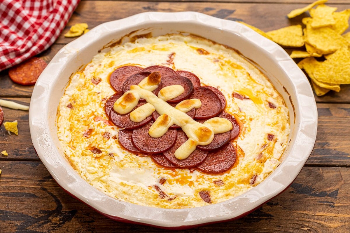 Pie plate with pepperoni pizza dip in it with pepperonis in a football shape.