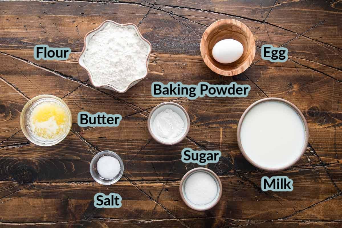 Ingredients Needed for recipe in bowls on a wood surface. Ingredients are flour, egg, baking powder, milk, sugar, salt, and butter