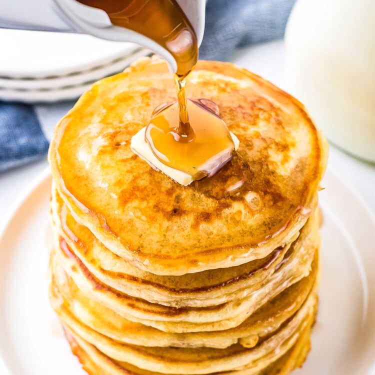 White plate with stack of pancakes and slab of butter on top with syrup being poured