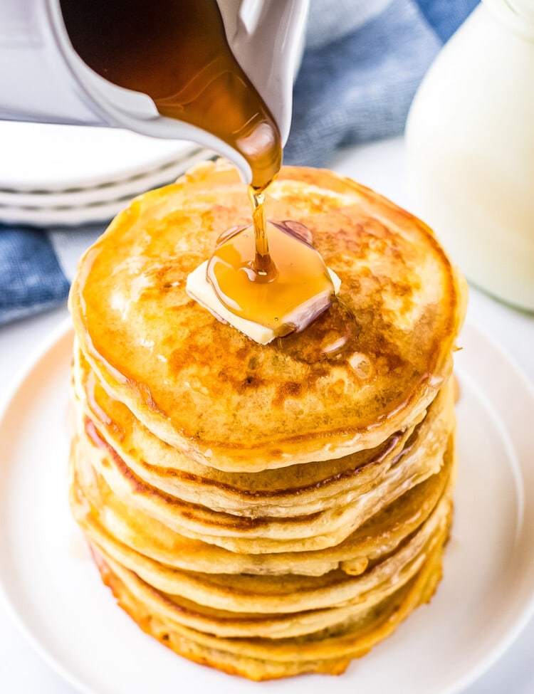 White plate with stack of pancakes and slab of butter on top with syrup being poured