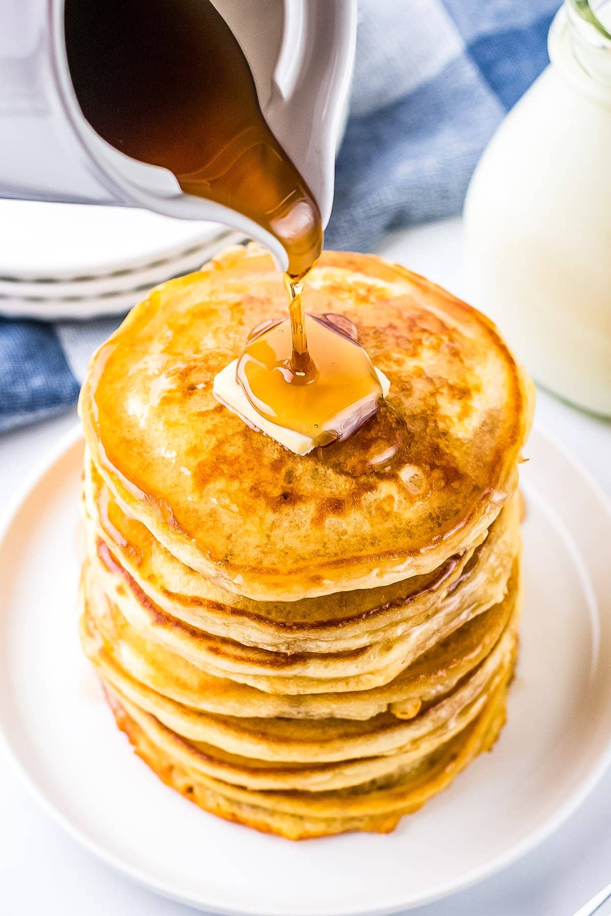 White plate with stack of pancakes and slab of butter on top with syrup being poured