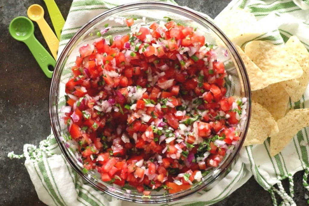 Overhead image of a glass bowl of pico de gallo with green and white napkin, chips and measuring spoons next to it