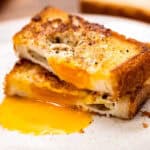A close-up of a toad in a hole, a slice of golden brown toasted bread with a fried egg cooked in the center. The egg yolk is runny and spilling onto the plate, with black pepper seasoning on top. The dish is served on a white plate with a blurred background.