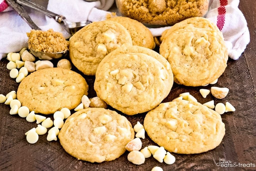 Brown board with White Chocolate Macadamia Nut Cookies laying on it and whtie chocolate chips plus brown sugar in teaspoon