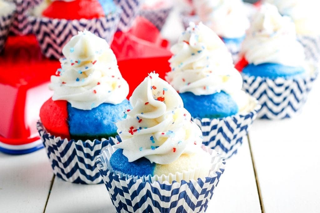 Multiple 4th of July Cupcakes with red white and blue cake in a chevron blue and white cupcake liner and white frosting with sprinkles on a white background