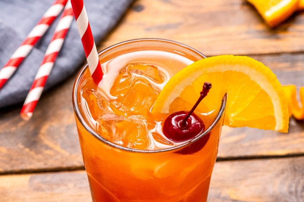 wood background with mixed cocktail garnished with cherry and orange slice and ride striped straw