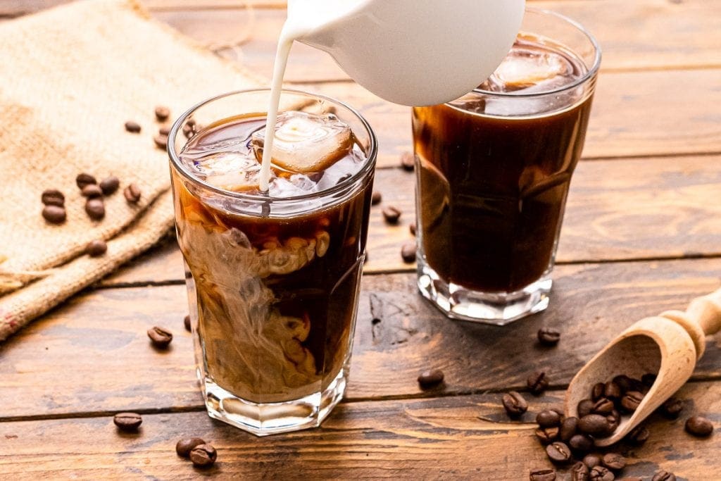 White pitcher pouring milk into a cold brew coffee in glass. Another glass of coffee behind that one on. On a wooden background with coffee beans on it.