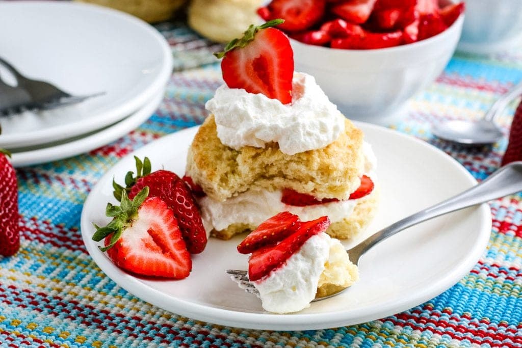 White plate with strawberry shortcake on it. A fork in front of it with a bite of dessert on it.