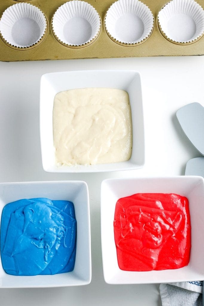 Overhead image with three square bowls one with white cake mix, one with red cake mix and one with blue cake mix. Muffin pan with white liners in it behind that. 