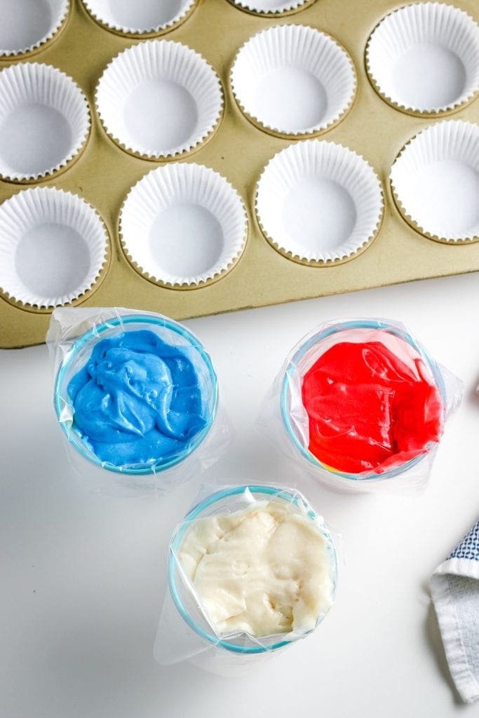 Overhead image cups holding bags of red, white and blue cake mix and muffin pan with white liners behind it.