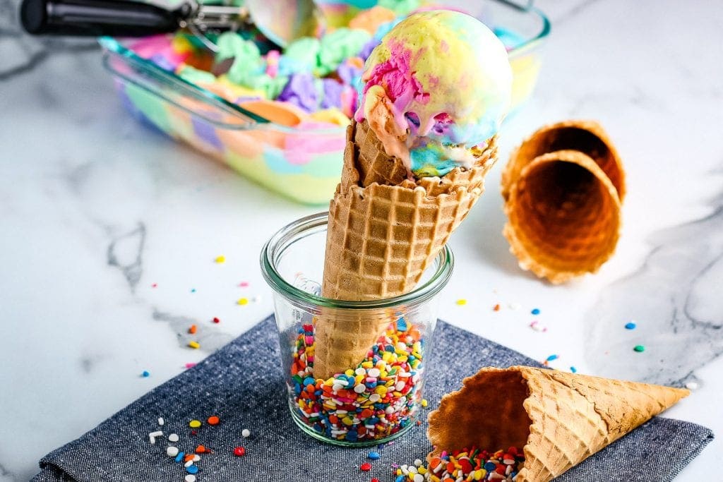 Waffle cone being held in glass jar that's topped with a scoop of rainbow ice cream. More waffle cones in background along with ice cream.