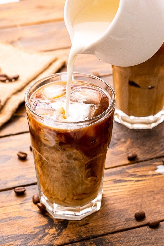 White pitcher pouring milk into a glass of cold brew coffee that's on a wooden background with coffee beans on it.