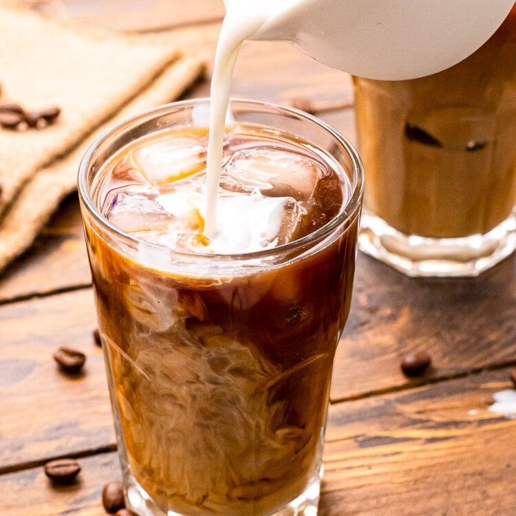 White pitcher pouring milk into a glass of cold brew coffee that's on a wooden background with coffee beans on it.