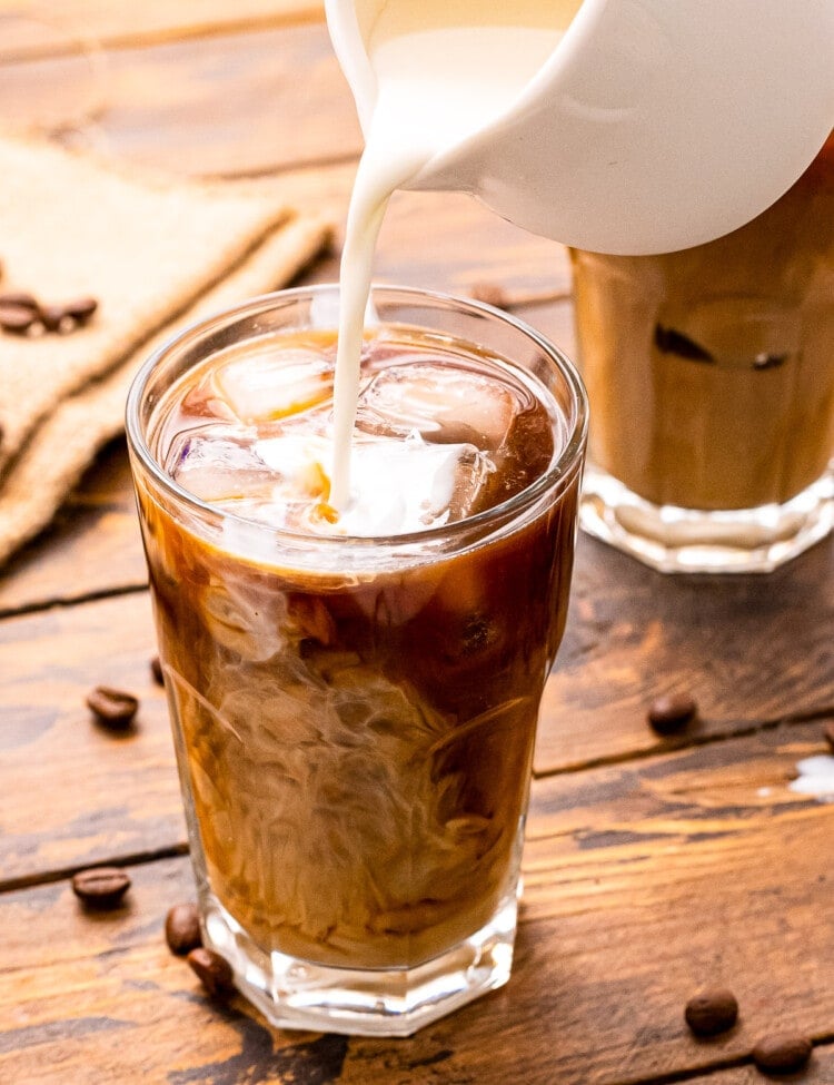 White pitcher pouring milk into a glass of cold brew coffee that's on a wooden background with coffee beans on it.