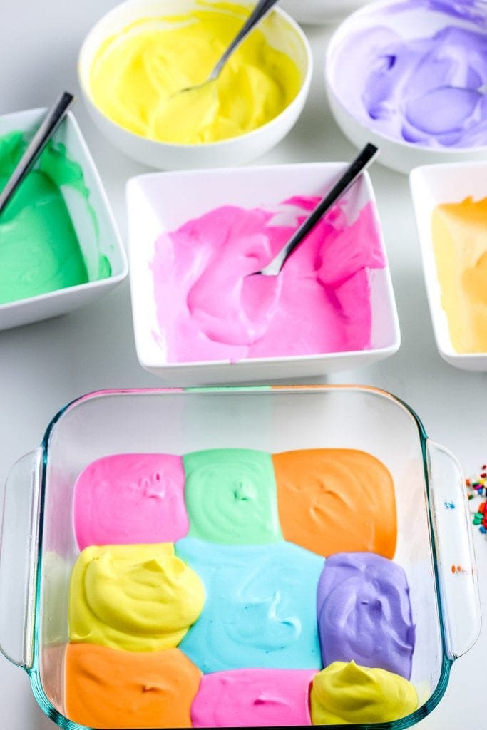 Glass baking dish with squares of different colors of ice cream and bowls of mixture behind it.