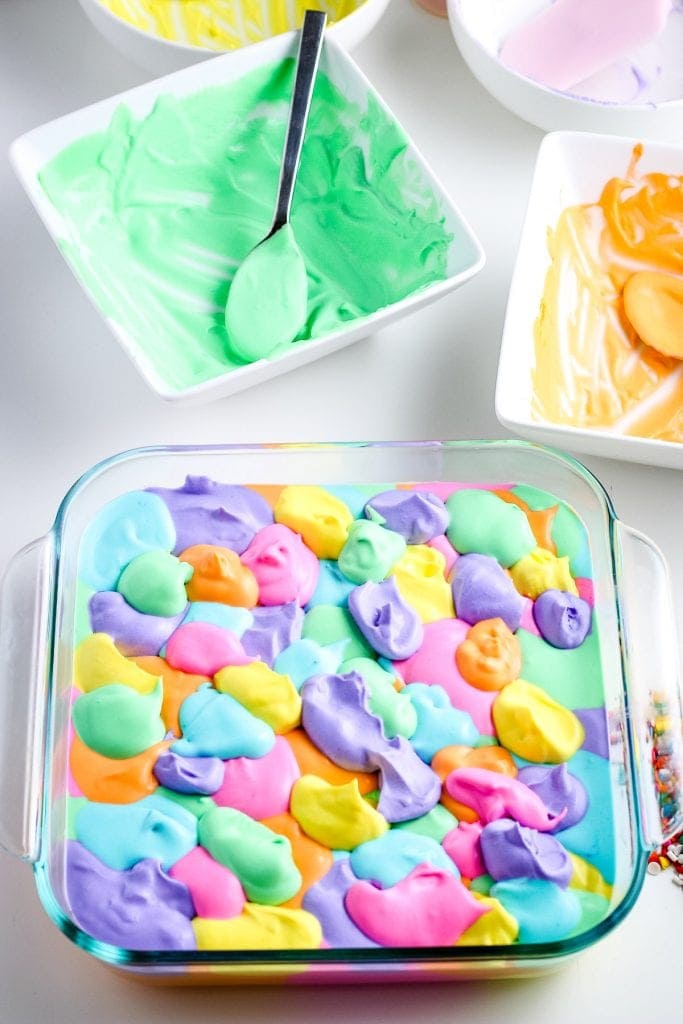 Glass baking dish with all the ice cream colors for rainbow ice cream in it and a green and orange bowl of ice cream mixture behind it.
