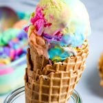 Waffle cone topped with rainbow ice cream being held by glass container.