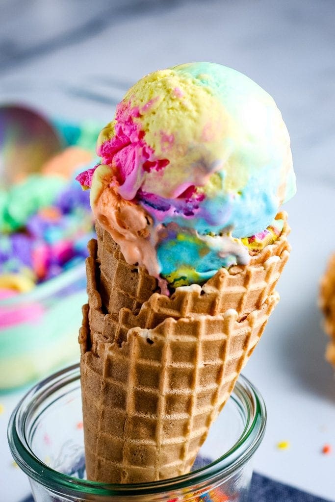 Waffle cone topped with rainbow ice cream being held by glass container.