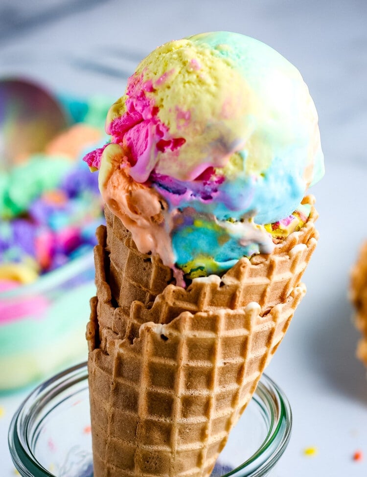 Waffle cone topped with rainbow ice cream being held by glass container.