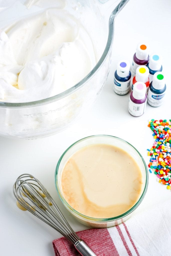 Glass bowl with sweetened condensed milk and vanilla mixed together, whipped cream in glass bowl behind it and food coloring containters.