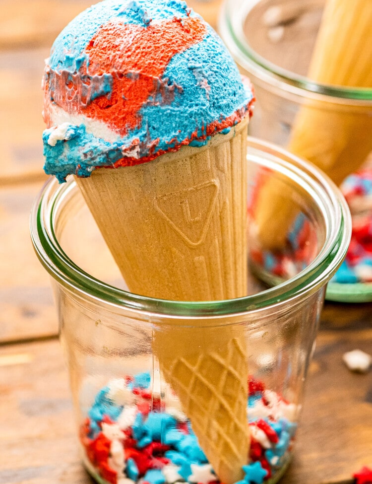 Glass jar holding a cone with red white and blue ice cream with sprinkles in the bottom of jar.
