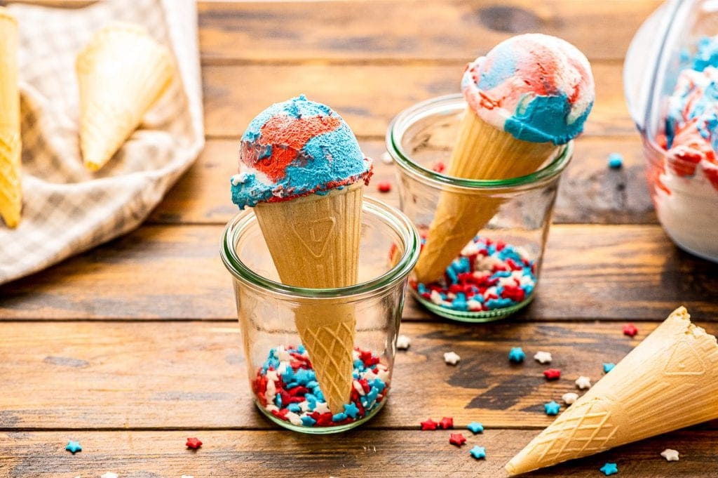 Two glass jars with sprinkles in bottom of jar and holding a cone of red white and blue ice cream. More sprinkles on wooden background and a cone laying down.