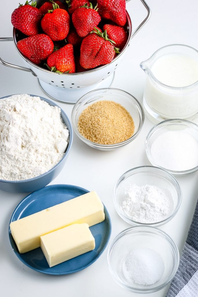 Ingredients needed to make Strawberry Shortcake in glass bowls and plates. Ingredients include butter, sugar, flour, strawberries, brown sugar, salt, milk and more.