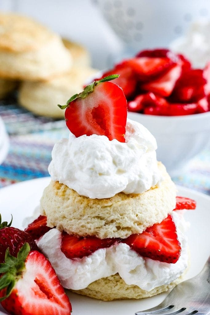 A strawberry shortcake on white plate with strawberries in background and biscuits.