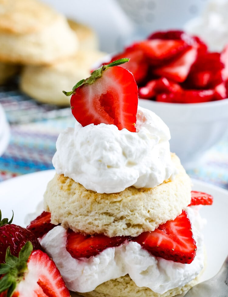 A strawberry shortcake on white plate with strawberries in background and biscuits.