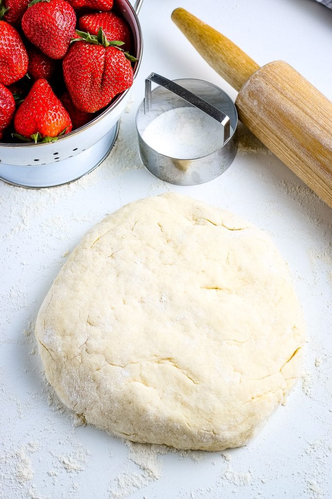 Biscuit dough before rolling out with strawberries in a bowl and wooden rolling pin behind it. 