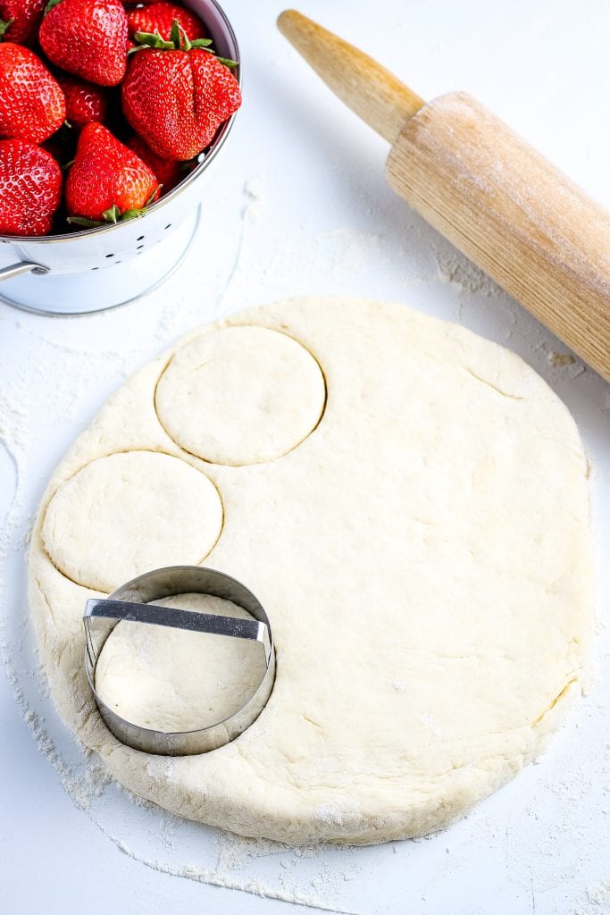 A circular rolled out biscuit dough with a biscuit cutter cutting biscuits. Fresh strawberries in a bowl and wooden rolling pin behind it.
