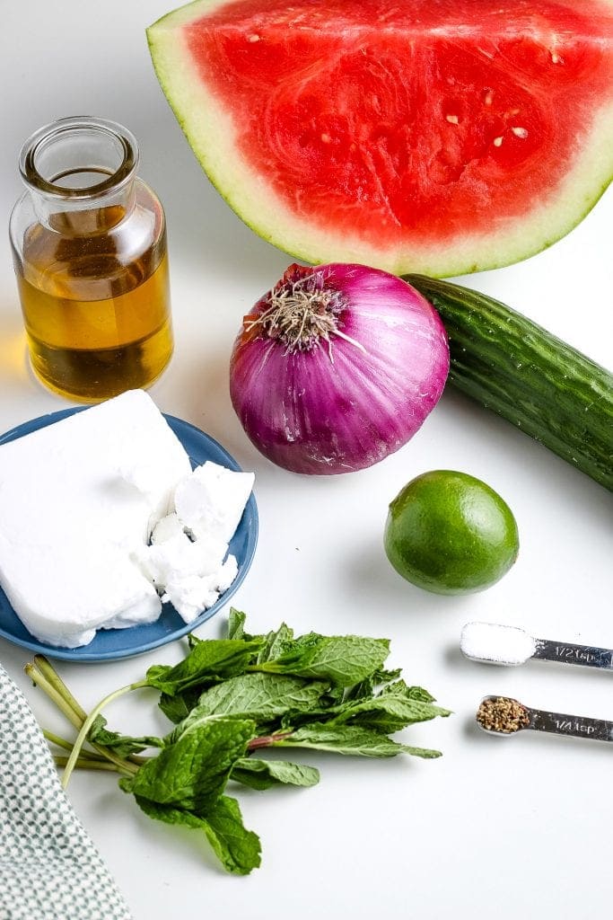 Portrait image showing ingredients including olive oil, feta cheese, red onion, watermelon, cucumber, mint and lime.