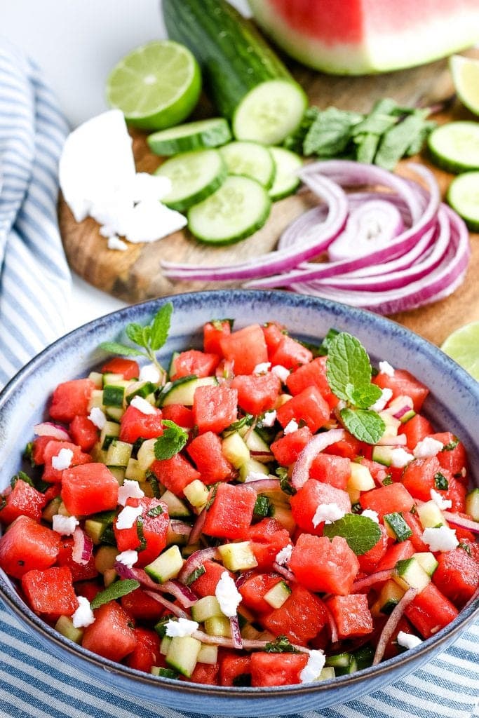 Portrait photo of watermelon salad in blue bowl with red onion, cucumber, feta cheese in background