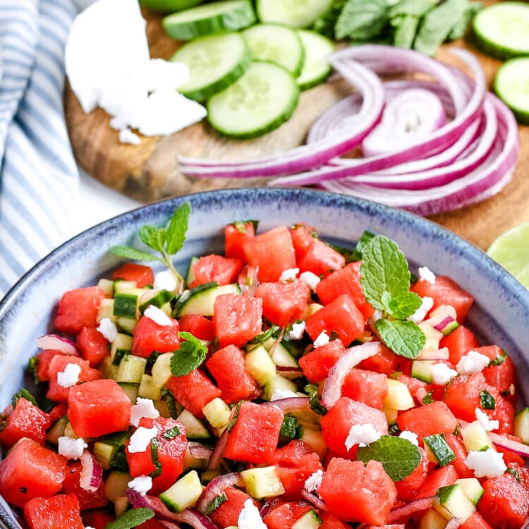 Portrait photo of watermelon salad in blue bowl with red onion, cucumber, feta cheese in background