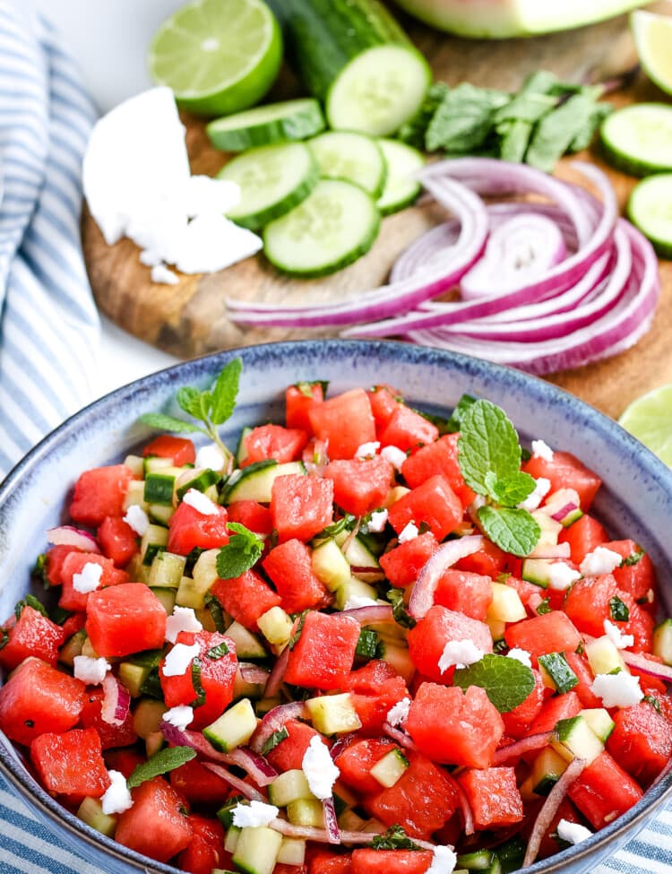 Portrait photo of watermelon salad in blue bowl with red onion, cucumber, feta cheese in background