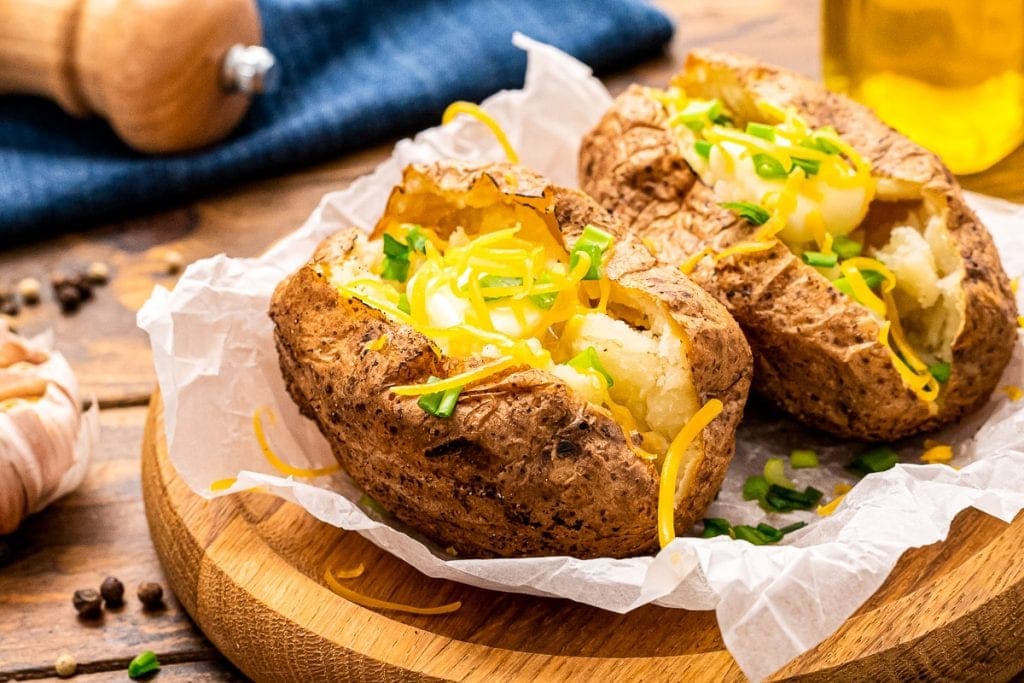 Wooden platter with baked potatoes lying on freezer paper. Baked Potatoes are cut open with cheese and green onions garnishing.
