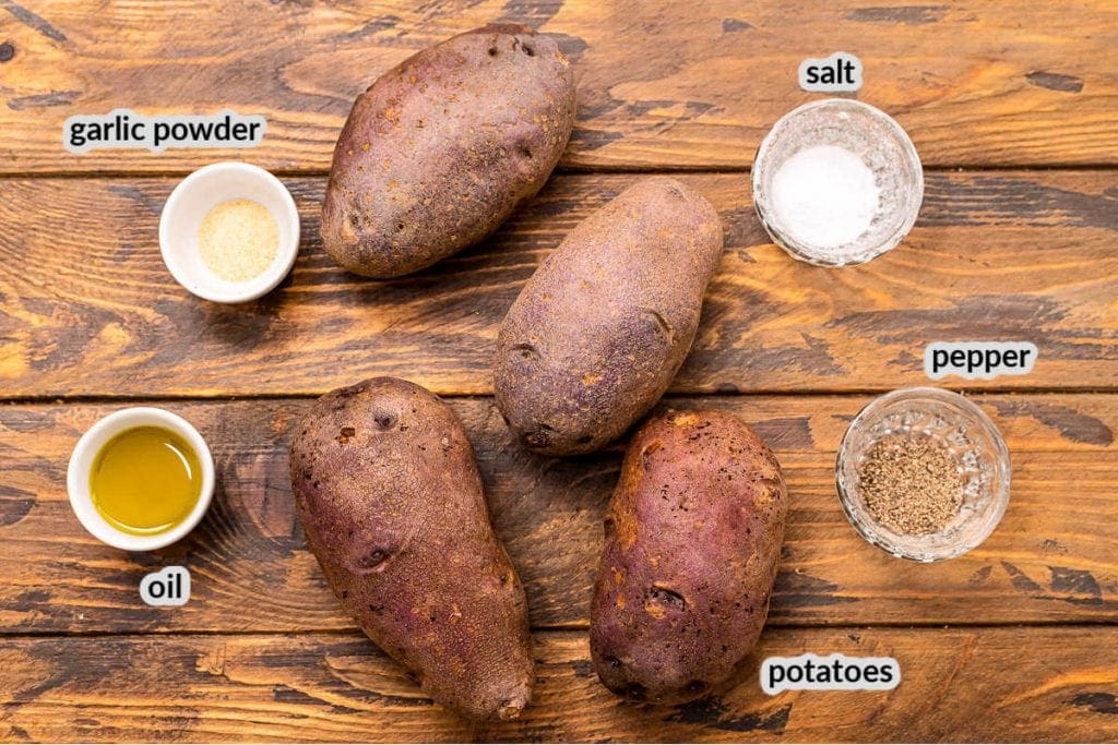 Overhead image showing wooden background with ingredients to make baked potatoes including salt, pepper, oil, garlic powder and potatoes