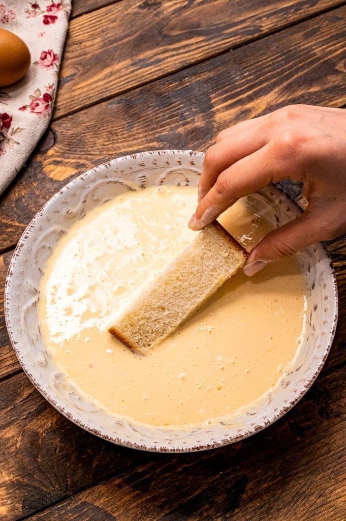 sliced stick of bread being dipped in custard mixture to make french toast sticks