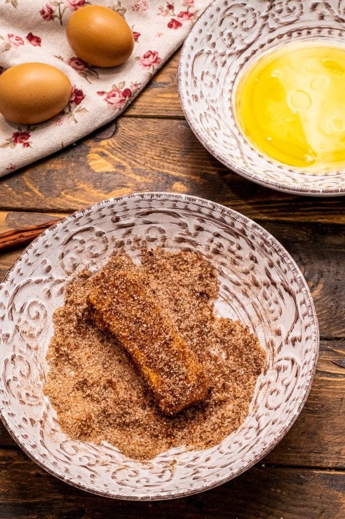 Bowl with cinnamon sugar in it and a french toast stick being rolled in it. A bowl of melted butter in background.