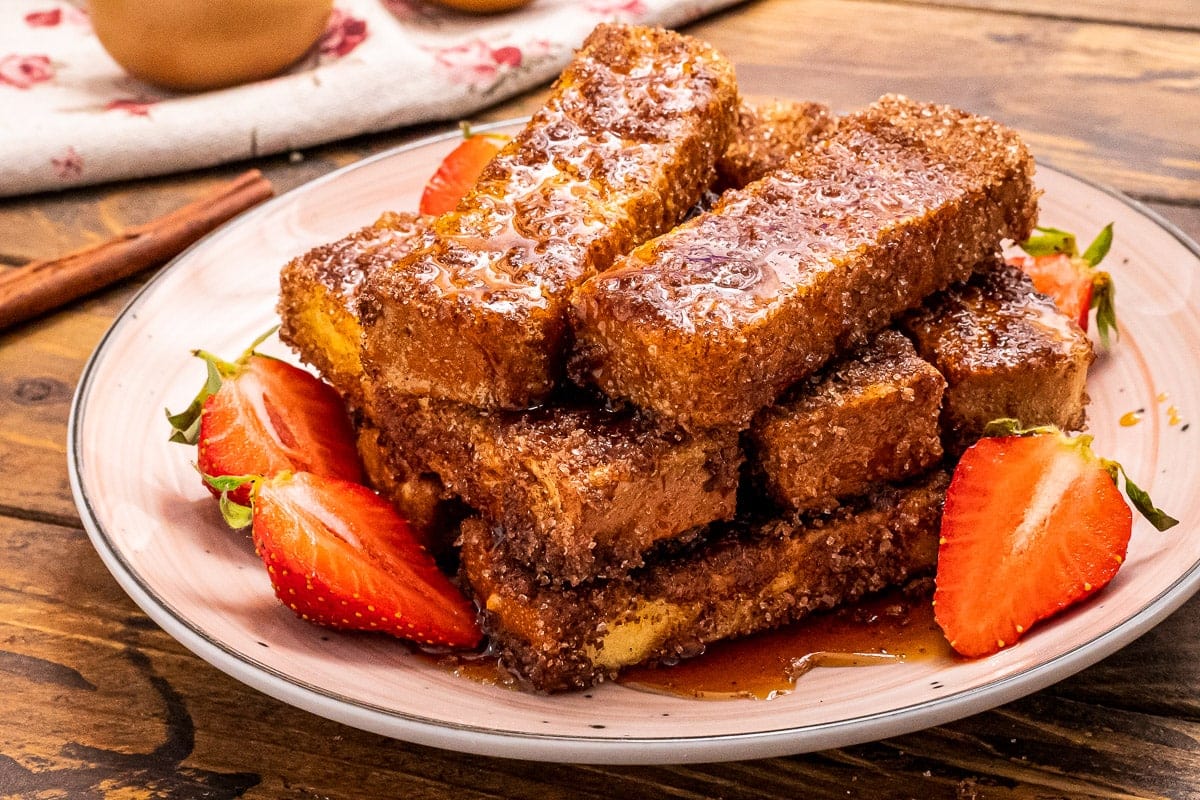 White Plate with French Toast sticks on it along with sliced strawberries beside it on a wood background
