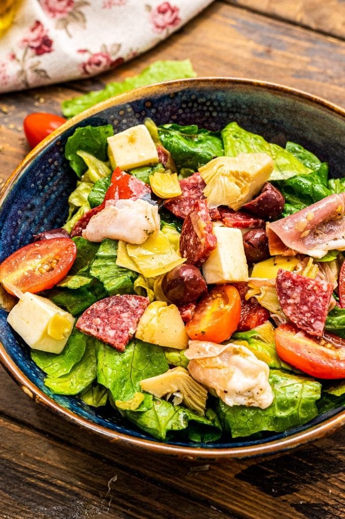 Portrait photo of a dark blue ceramic bowl with tossed Antipasto Salad in it on a wooden background